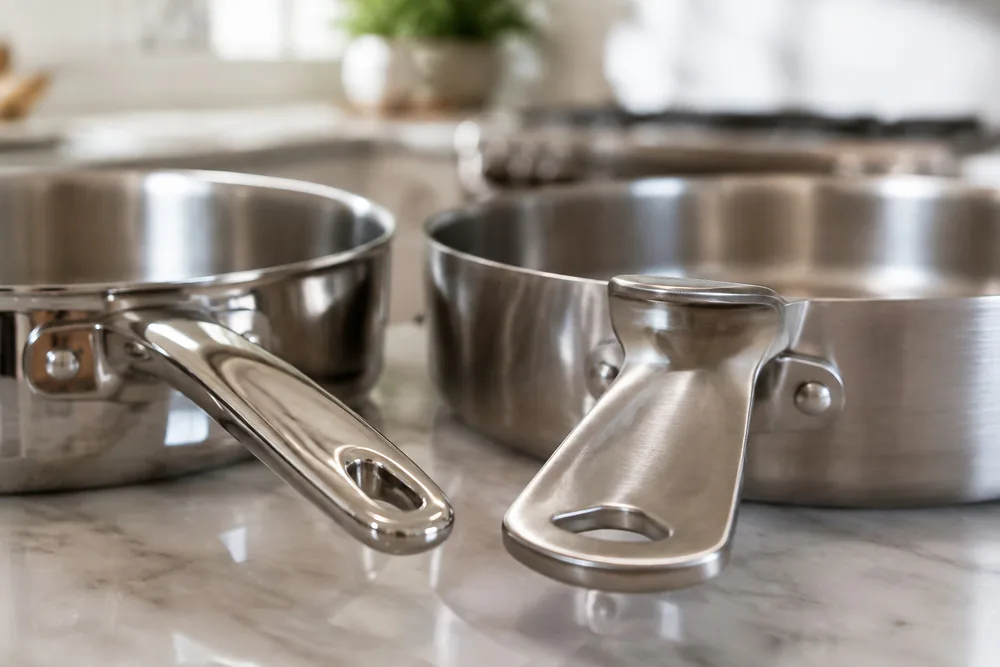 Close-up of an All-Clad D3 polished handle beside an oversized D5 brushed handle with a finger and thumb stop.