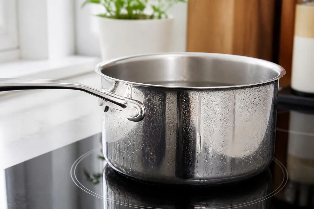 An All-Clad D3 stainless saucepan on a glass induction cooktop, water condensing on the polished exterior.