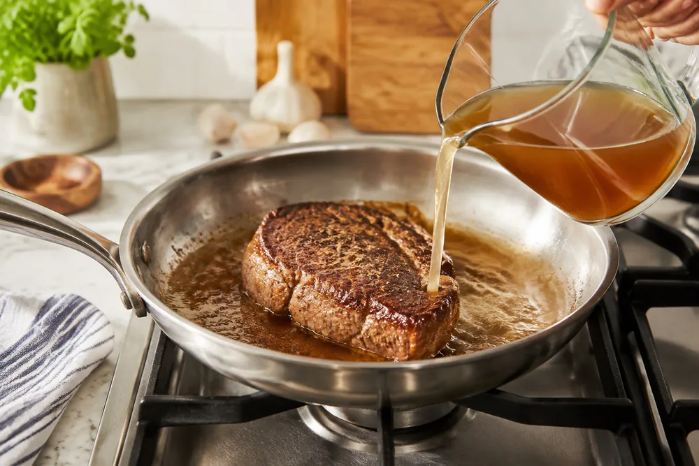 A steak searing in an All-Clad D3 12-inch fry pan as broth is poured in to lift the browned fond.