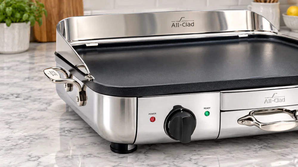 Close-up of the All-Clad 99014GT electric griddle showing the cast stainless handle, polished exterior and indicator lights on a kitchen counter