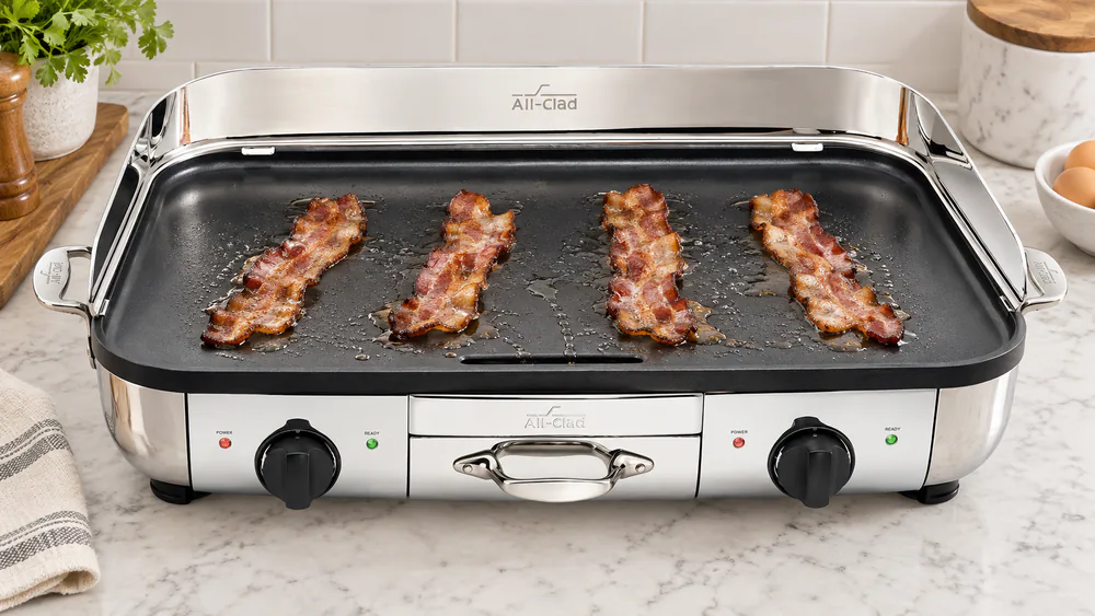 Overhead view of the All-Clad 99014GT cooking plate with bacon strips laid across it and grease running toward the central drain slot