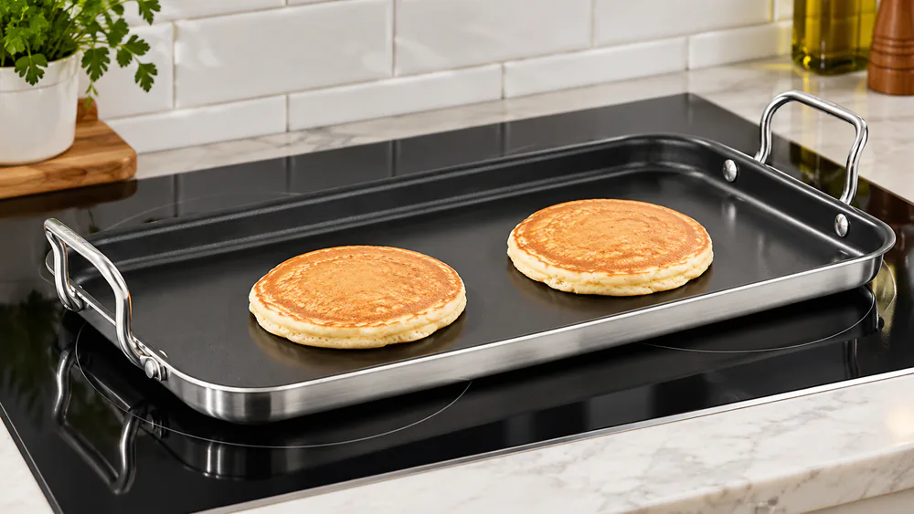 A Cuisinart MultiClad Pro double burner griddle with brushed stainless exterior on an induction cooktop with two pancakes cooking