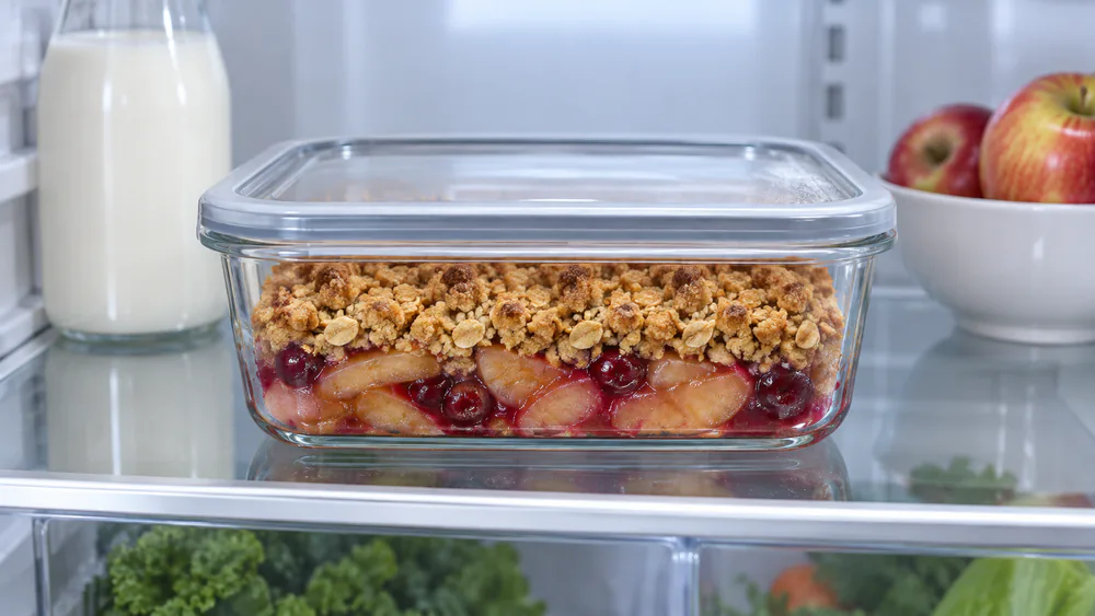 Leftover apple cranberry crisp stored in a clear airtight glass container in the refrigerator, oat topping still intact on top of the fruit.
