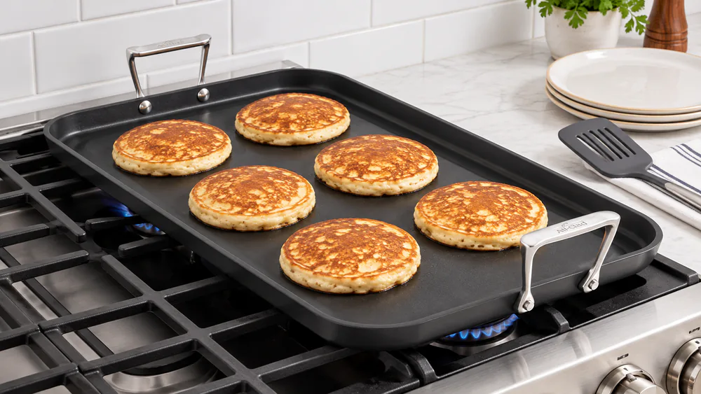 An All-Clad HA1 13x20 hard-anodized double burner griddle on a gas range with a row of golden pancakes cooking across its dark matte surface