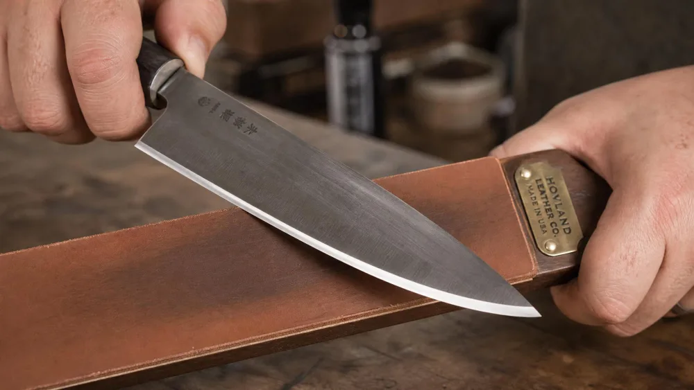 A knife being stropped on a leather strop, edge trailing and a light micro-bevel polish visible.