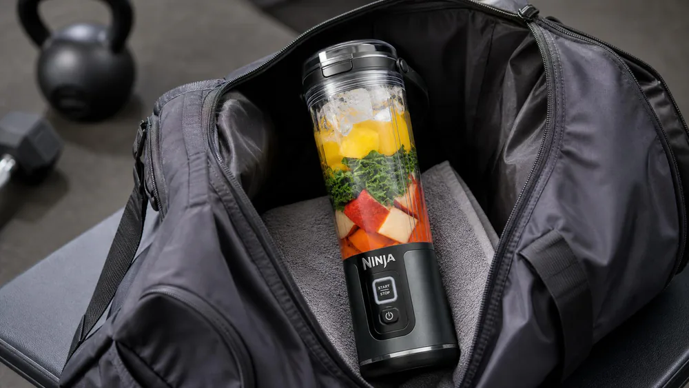 A Ninja Blast cordless blender with its 18-oz vessel sitting inside a gym bag next to a folded towel, ready to carry.