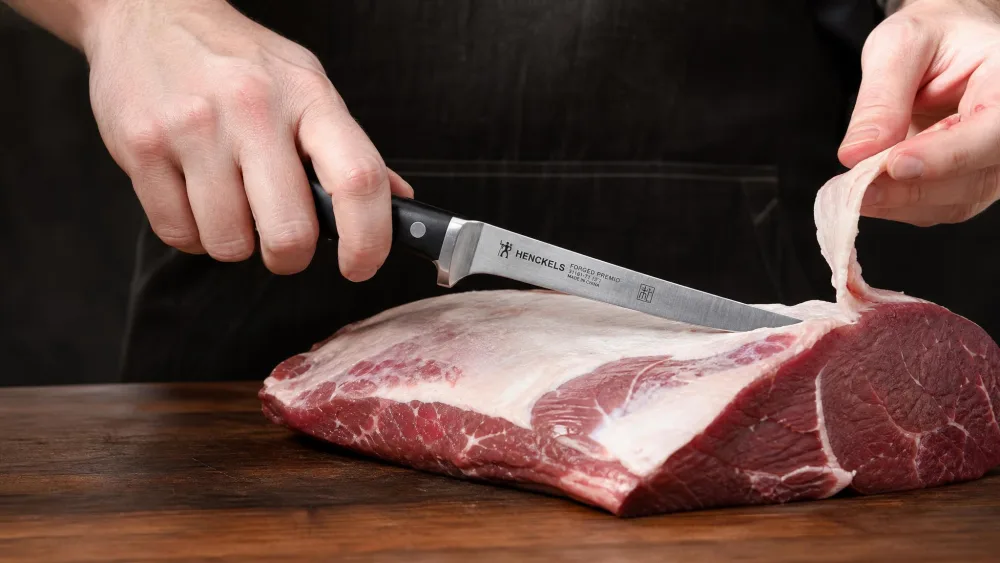A HENCKELS Forged Premio boning knife with a triple-rivet handle trimming fat from a large cut of beef.