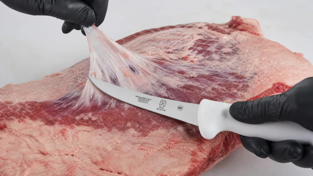 A Mercer Ultimate White curved boning knife trimming silverskin from a brisket, its white handle visible.