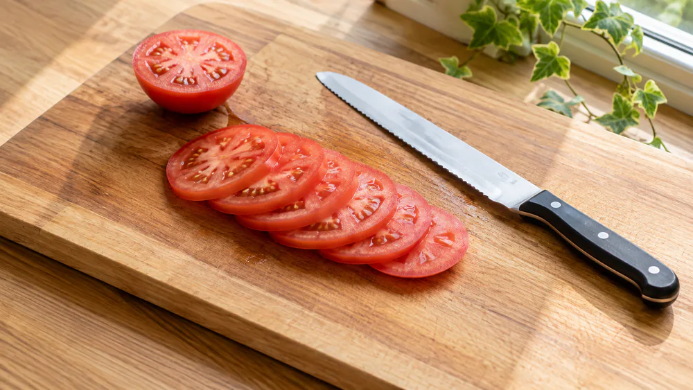 A ripe tomato cut into thin, unbroken rounds on a cutting board beside the bread knife that sliced them, from the tomato test.
