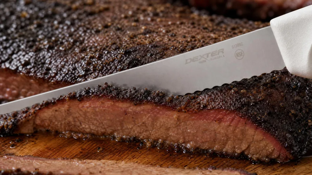 Close-up of the Dexter-Russell scalloped slicer cutting through the dark bark of a smoked brisket.