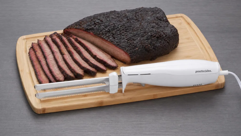 The Proctor-Silex electric knife with two reciprocating stainless steel blades assembled on a cutting board beside a brisket.