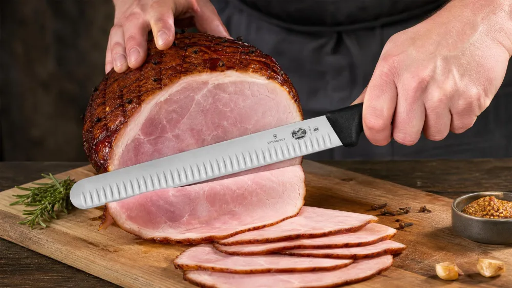 The Victorinox Fibrox Pro 12-inch Granton slicer slicing a smoked ham, hollow-edge dimples visible along the blade.