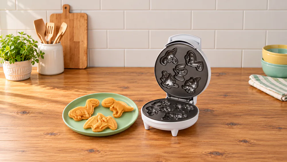 Three small 3D dinosaur waffles on a toddler plate next to the Waffle Wow dinosaur mini waffle maker