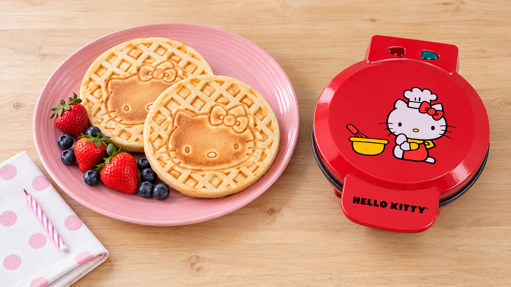 Two Hello Kitty waffles on a pink plate beside the red Hello Kitty waffle maker on a birthday breakfast table