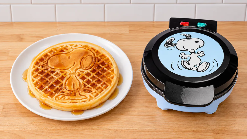 A round Snoopy-imprinted waffle with syrup on a plate next to the closed Peanuts waffle maker with Snoopy on the lid