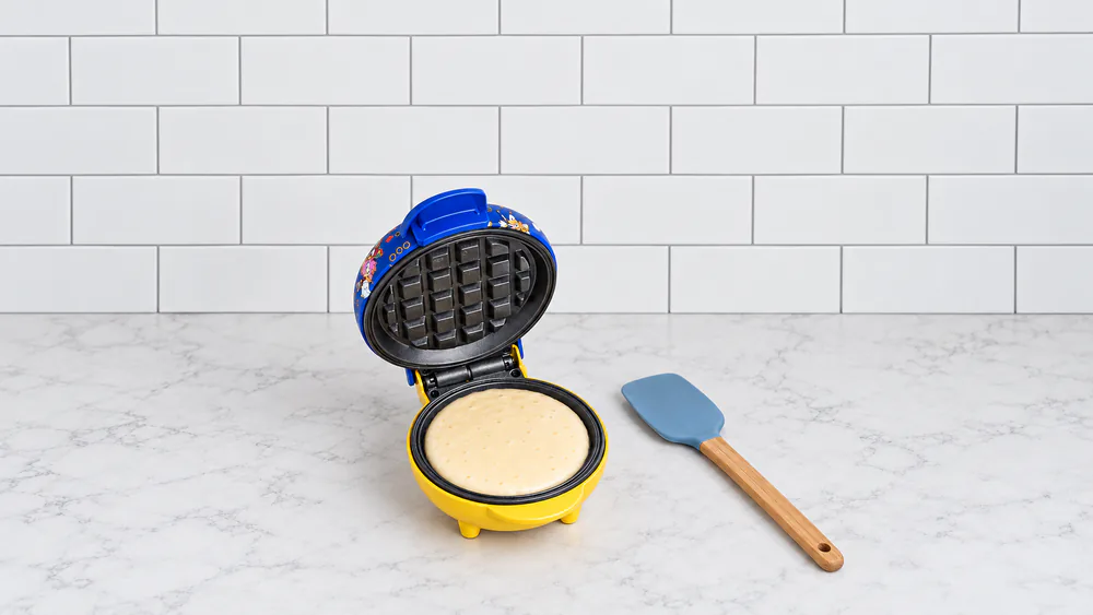 The Sonic the Hedgehog mini waffle maker open on a counter with batter poured into the plate and a spatula beside it