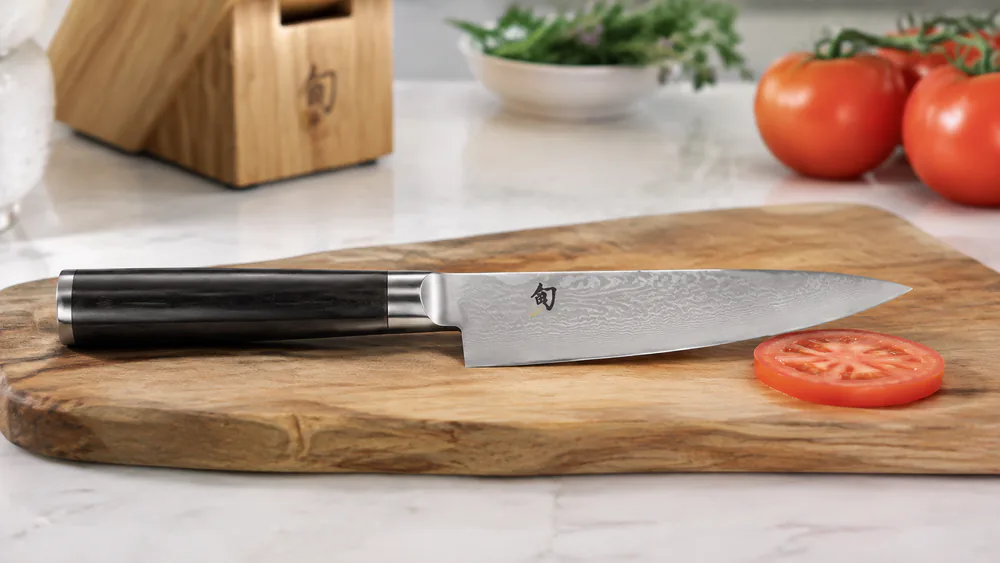 Close-up of the Shun Classic chef's knife edge, Damascus layering visible on the blade face, a tomato slice beside it.