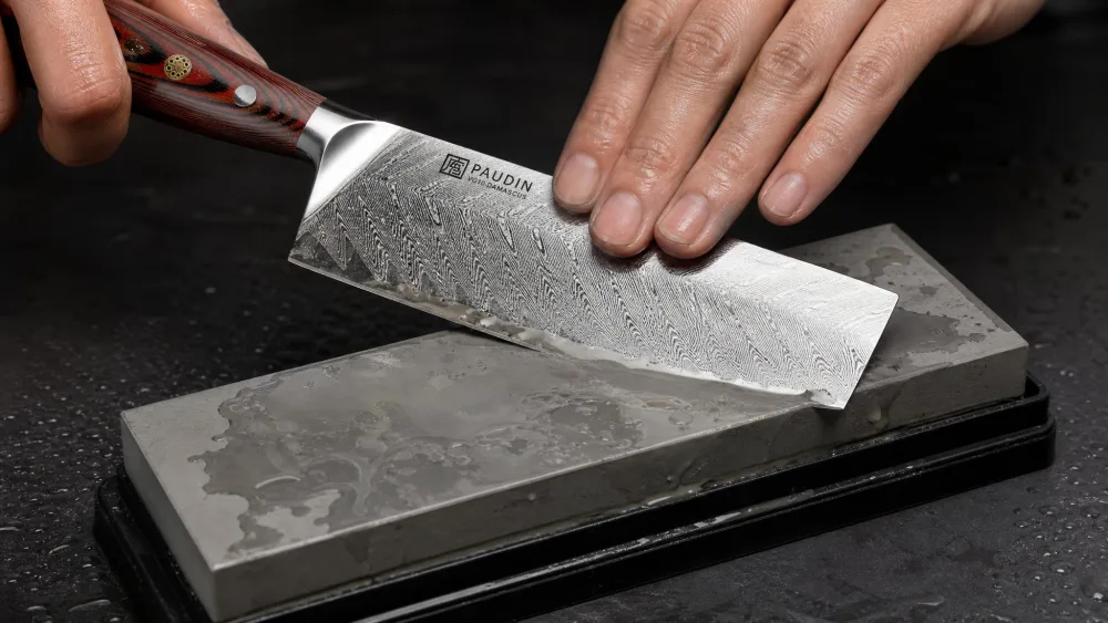A cleaver being sharpened on a whetstone with water and steel slurry on the stone surface, the blade held at a consistent angle.