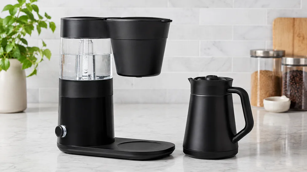 An OXO Brew 9-Cup Coffee Maker on a kitchen counter with its double-wall thermal carafe beside it, the single control dial visible in profile.