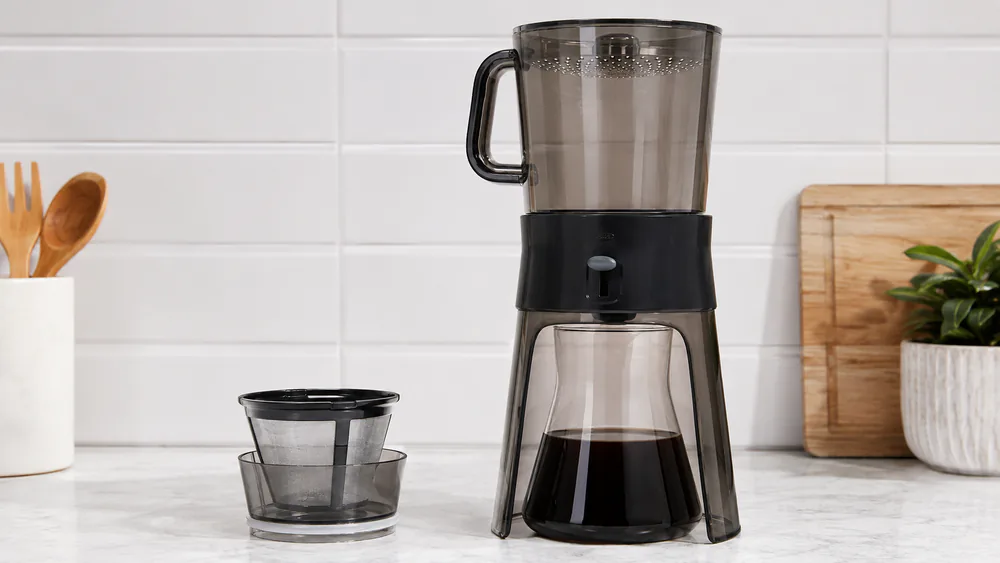 An OXO Good Grips Cold Brew carafe full of dark concentrate on a counter, the brew-release switch in the open position and the mesh filter basket beside it.