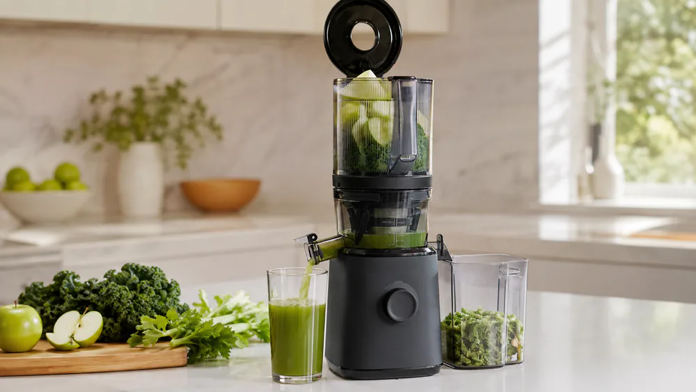 A Nama J2 Cold Press Juicer with its self-feeding hopper loaded with apples and a tall glass of freshly pressed juice beside it.