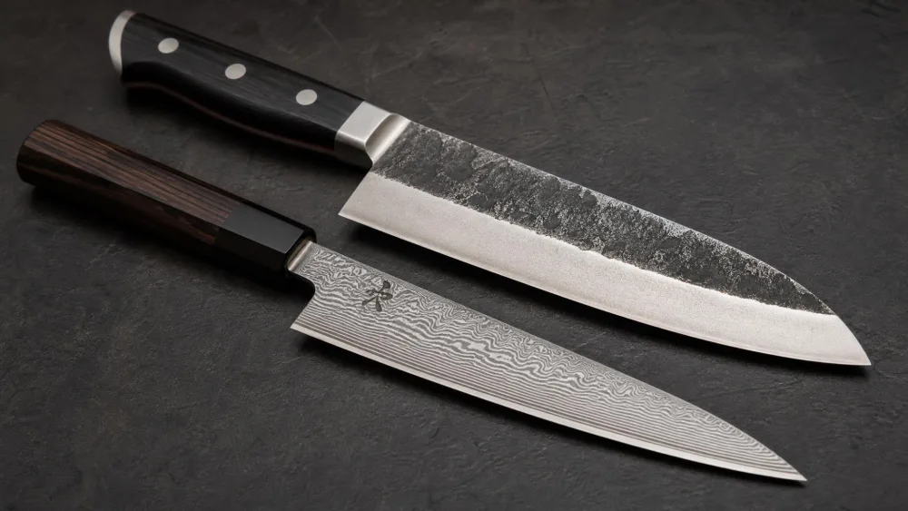 A forged German chef's knife with a thick spine and full bolster resting beside a slimmer Damascus-clad Japanese chef's knife, both angled to show the difference in blade profile and bevel.