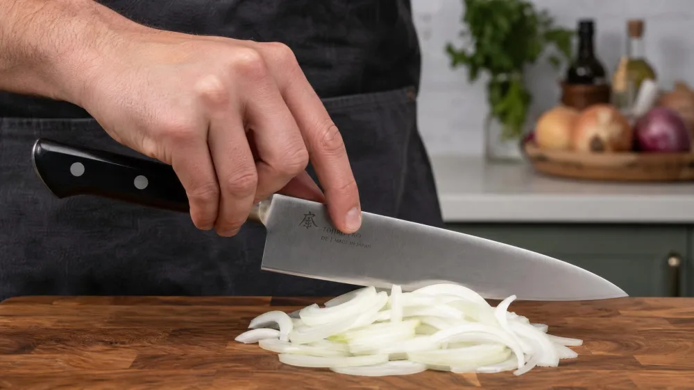 A hand holding an 8-inch chef's knife in a pinch grip, thumb and forefinger gripping the blade just ahead of the bolster, mid-cut over a pile of sliced onion.