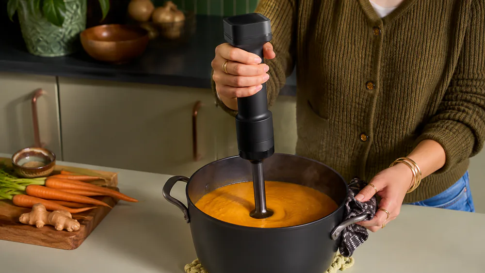 A KitchenAid Go cordless hand blender pureeing a pot of hot soup, held one-handed with no cord in sight.