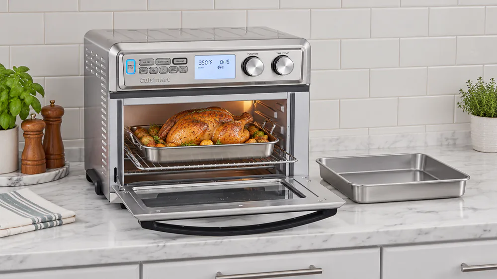A whole roast chicken resting in the wide cavity of a Cuisinart TOA-95 digital toaster oven next to a 9 by 13 baking pan.