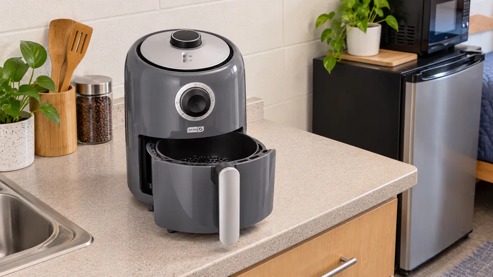 Dash Compact 2-Qt air fryer on a small dorm room counter beside a mini fridge with the nonstick basket half pulled out