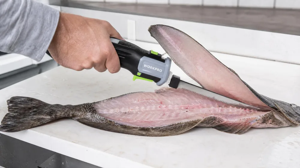A hand guiding an electric fillet knife down a fish backbone at a cleaning table, a neat fillet lifting away with the skin still attached.