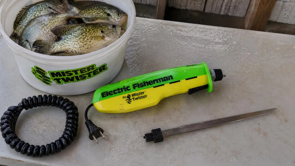 A Mister Twister 120V electric knife at a cleaning table beside a bucket of crappie and bluegill, serrated blade removed and resting next to it.