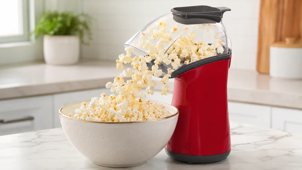 A Presto PopLite 04863 air popper running with the clear chute directing popcorn into a waiting bowl, the butter-melting cap resting on top.
