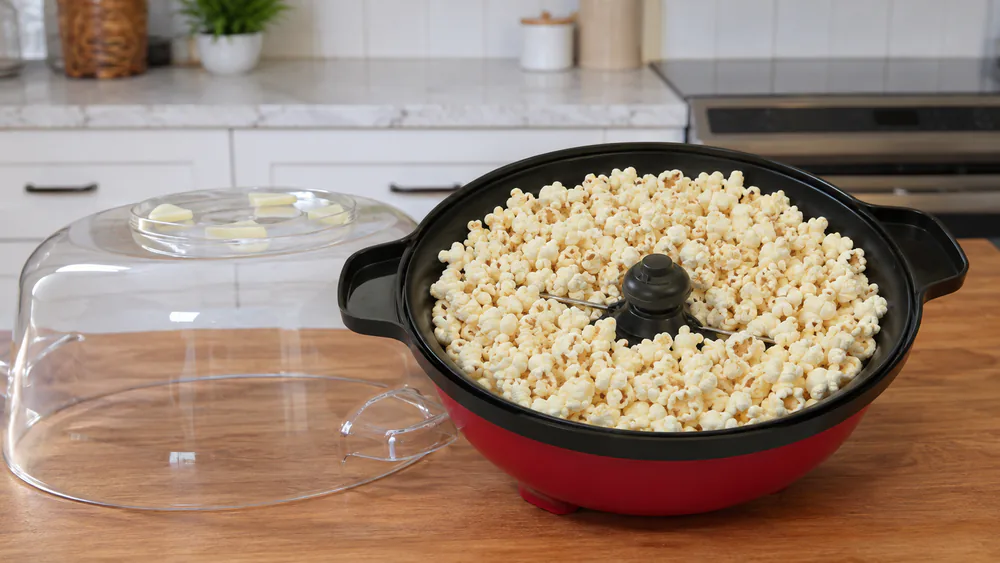 A West Bend Stir Crazy 82505 with its clear cover lifted, a full 6-quart batch of popped popcorn inside, the motorized stir rod visible beneath the kernels.