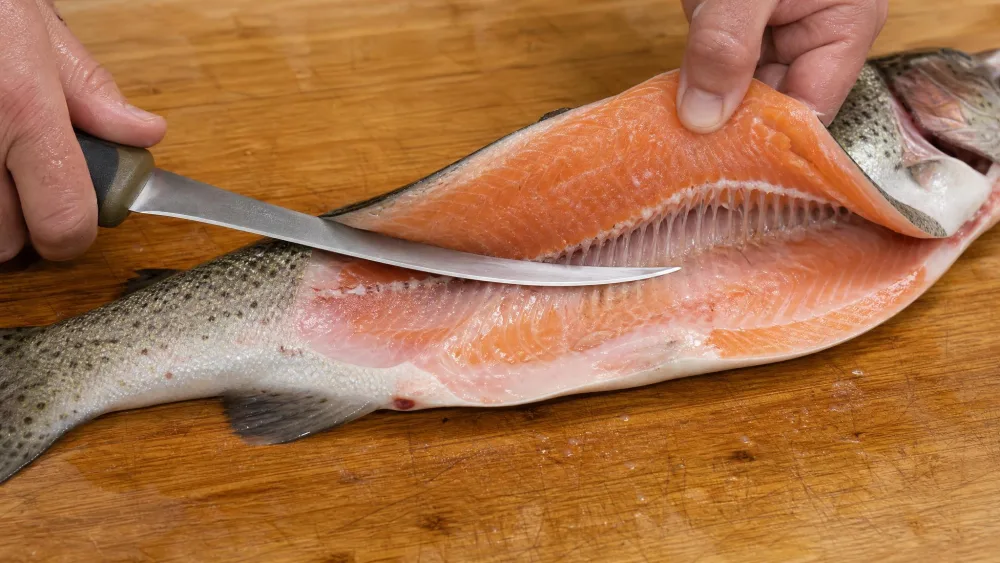 A fillet knife slicing a trout fillet off the backbone on a wooden cutting board, the blade flexing to follow the ribcage.