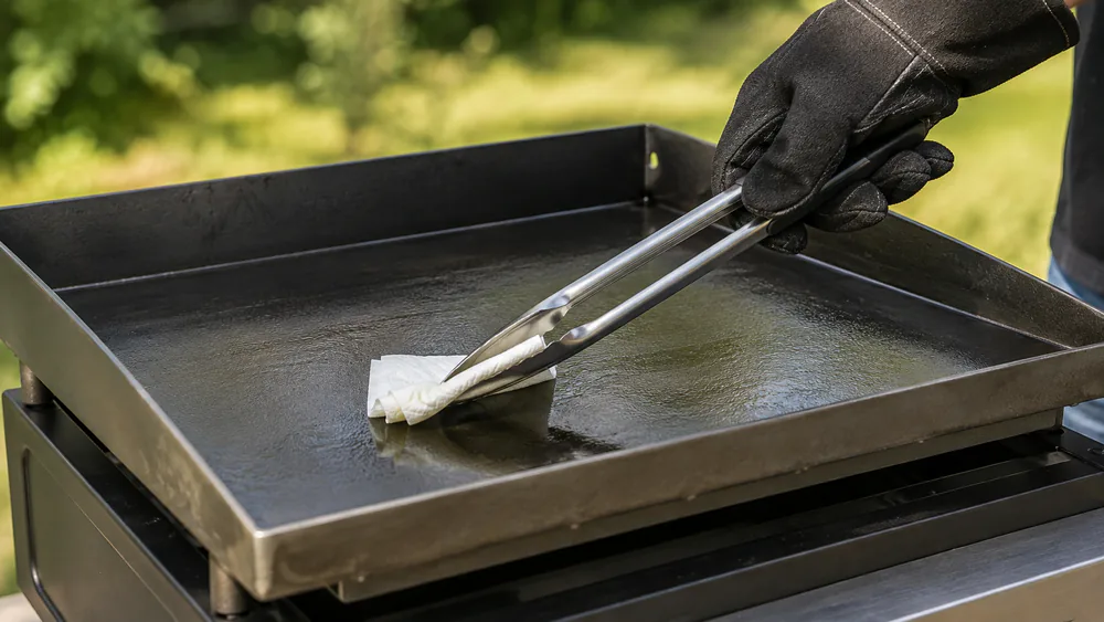 A hand spreading a thin layer of high-smoke-point oil across a warm carbon steel griddle cooktop with a paper towel.