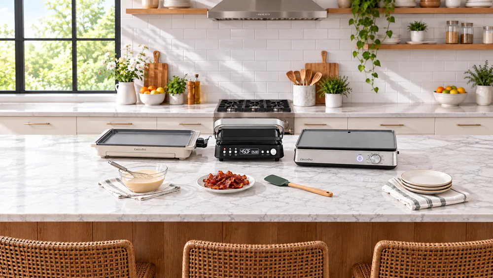 Three GreenPan electric griddles lined up on a kitchen counter with pancake batter, a plate of bacon, and a silicone spatula nearby