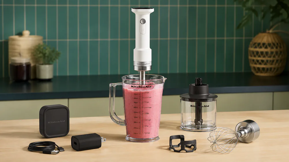 A KitchenAid Two-Speed hand blender standing in a tall glass of strawberry smoothie on a counter, ice visible at the base of the glass.