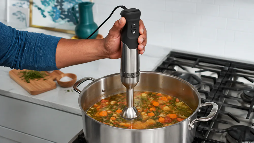 The Vitamix 5-Speed immersed in a large pot of vegetable soup, the coated bell guard visible against the side of the pot.