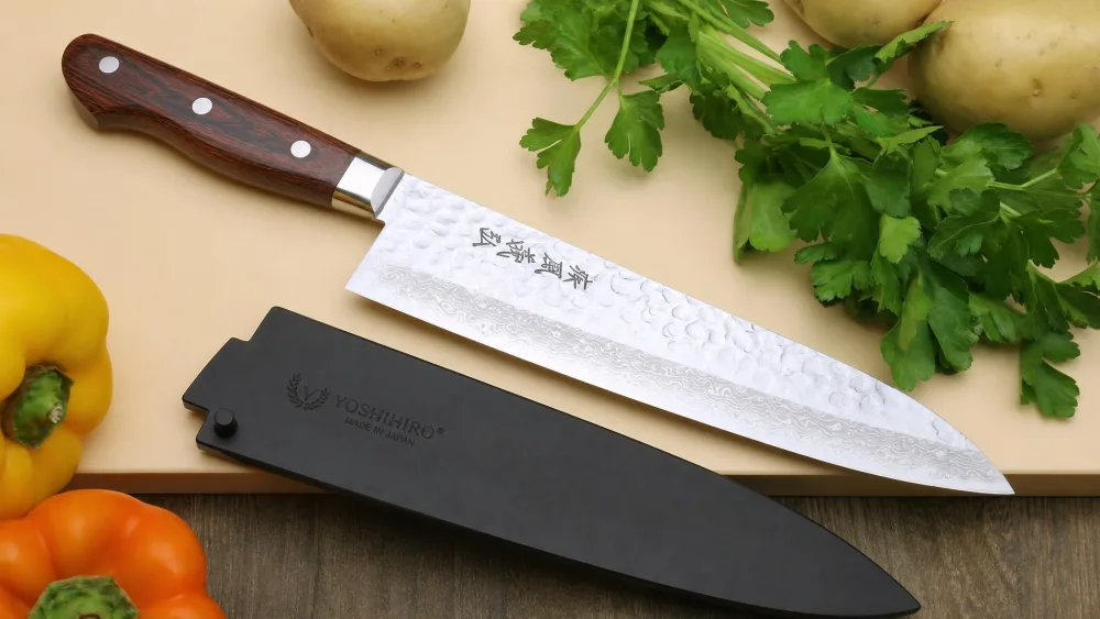 A Yoshihiro hammered Damascus gyuto with its Western mahogany handle laid beside a protective blade guard.