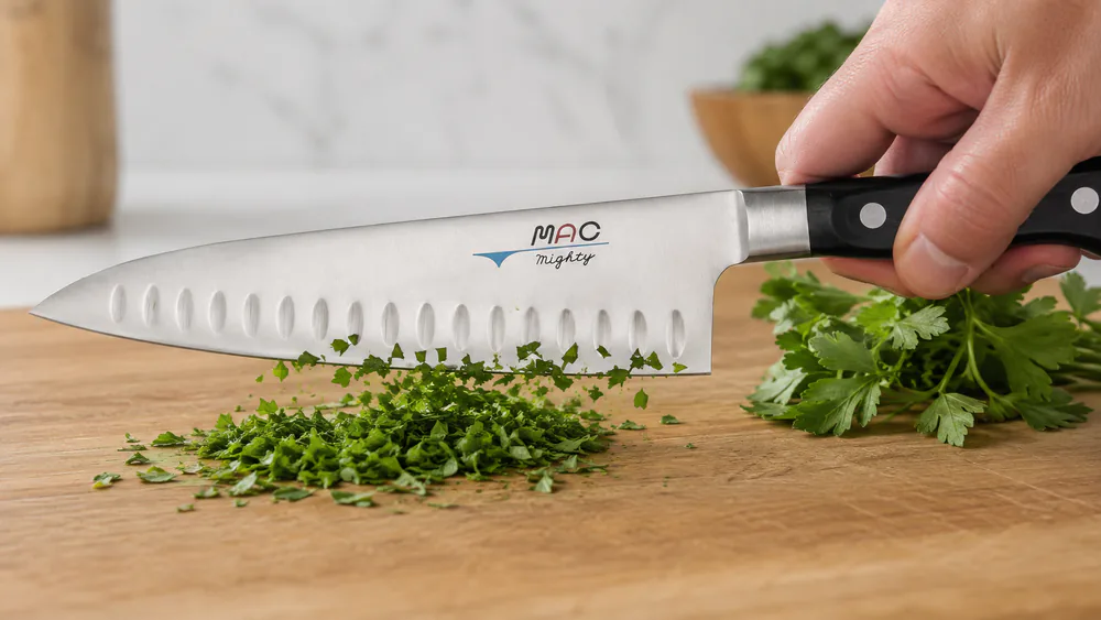 A MAC MTH-80 chef's knife with a dimpled blade chopping fresh herbs on a wooden cutting board, thin slices falling cleanly.