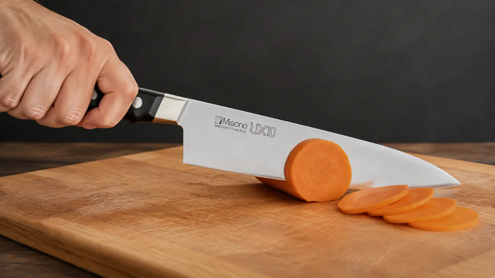 A Misono UX10 gyuto slicing through a dense raw carrot on a cutting board, the thin blade passing cleanly without wedging.