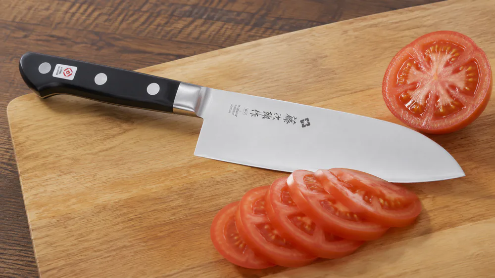 A Tojiro DP santoku with a black laminated handle slicing a ripe tomato into thin even slices on a wooden board.