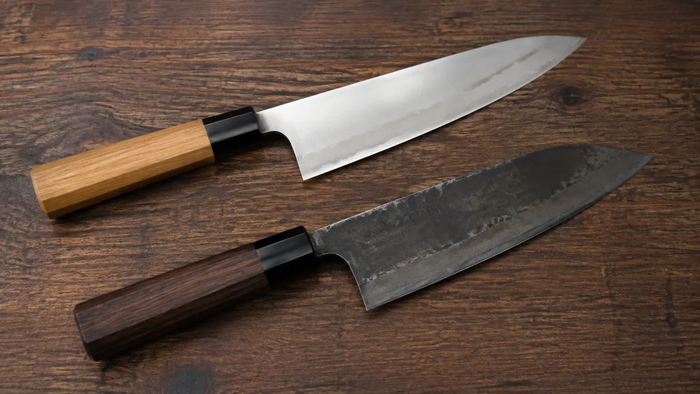 Two Japanese knife blades side by side, one stainless VG-10 with a bright finish and one high-carbon blade with a darker patina.