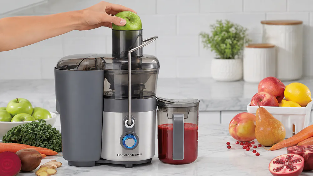 A whole apple going into the 3-inch Big Mouth chute of the Hamilton Beach Easy Clean juice extractor on a kitchen counter.