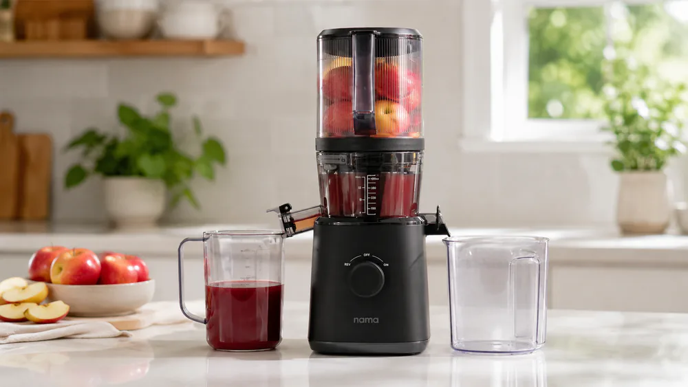 The Nama J2 cold press juicer with its hopper loaded with whole apples, showing the hands-free self-feeding chamber on a kitchen counter.