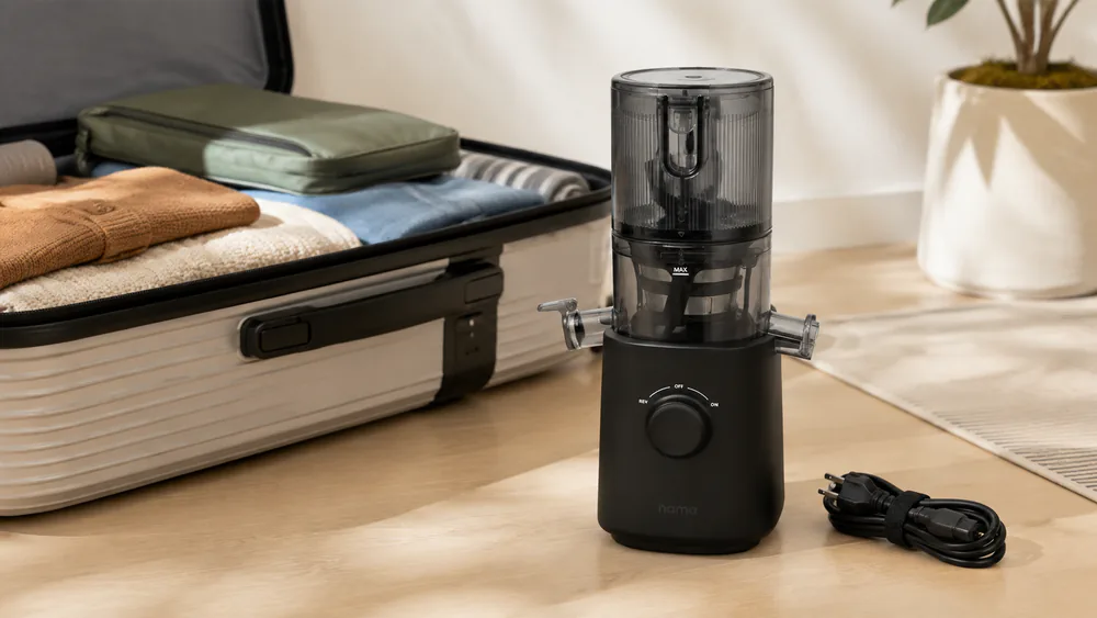 The compact Nama J3 cold press juicer beside a packed suitcase, showing its small footprint and detachable cord.