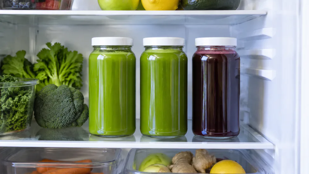 Two glass jars of bright green juice in a refrigerator, filled to the top, with a third jar of darker juice beside them.