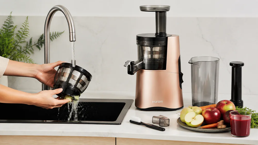 The wide filter basket and strainer of a Hurom H-AA slow juicer under a running kitchen tap, pulp rinsing off the mesh.
