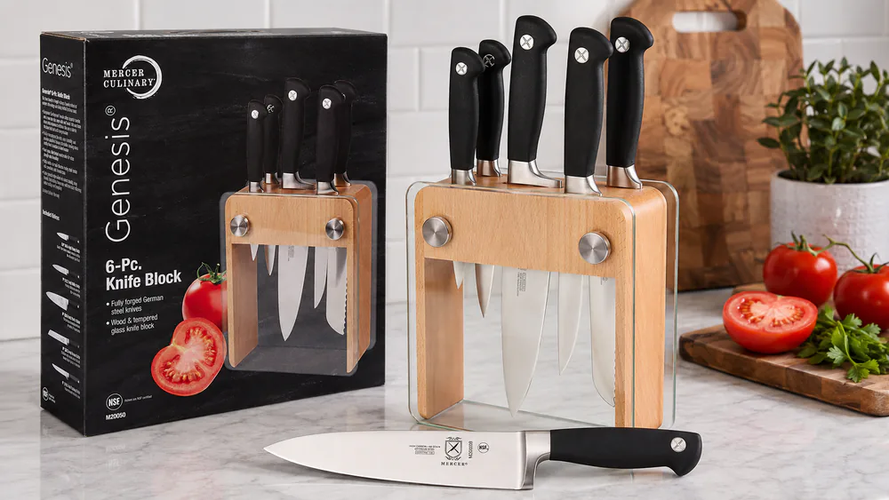 The Mercer Genesis 6-Piece set in its beechwood and tempered-glass block on a kitchen counter, chef's knife in the foreground.